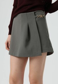 Jupe grise plissée avec un accent de chaîne dorée sur le côté et une texture lisse, présentant un design épuré et ajusté ainsi qu'une silhouette légèrement évasée.