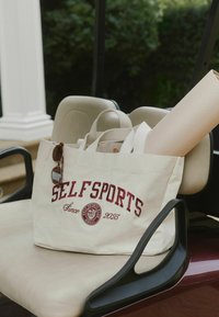 Borsa tote in tela beige con la scritta "SELFSPORTS" e un emblema color granata. Include occhiali da sole posati sulla borsa, sistemata sul sedile di un carrello da golf.