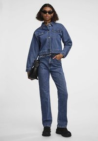 Jeans jakna s krojenjem iznad struka, plave boje, s kopčanjem na dugmad. Uparena s plavim trapericama ravnog kroja i crnim robusnim tenisicama. Crna torba.