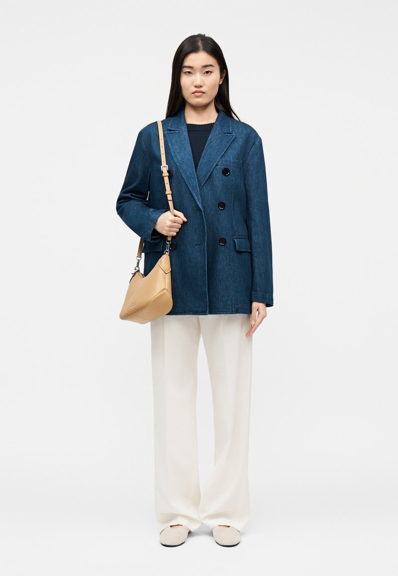 Γυναίκα που φοράει σκούρο μπλε διπλόμπουτο σακάκι, άσπρο παντελόνι με φαρδιά μπατζάκια, μπεζ παπούτσια και κρατάει μια μπεζ τσάντα ώμου.