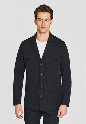 Młody mężczyzna w czarnej zapinanej na guziki kurtce z czterema przednimi kieszeniami, na białej koszuli, stoi na jednolitym jasnym tle.