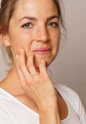 Gold ring with multiple clear gemstones set in a wavy pattern, worn on a hand touching the face. The background is neutral and simple.