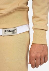 Felpa beige con polsini a coste, caratterizzata da un'etichetta visibile con la scritta "Reichstadt" e sottili dettagli ricamati. Texture morbida e design aderente.