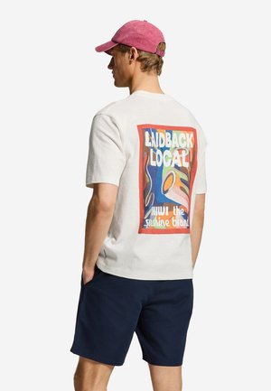 Mand iført en hvid t-shirt med en farverig "Laidback Local" grafik på ryggen, marineblå shorts og en lyserød baseballkasket, vendt væk.
