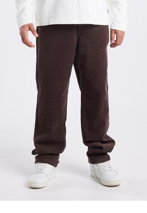 Bruine corduroy broek met verticale ribbels, losse pasvorm en een lichte uitlopende zoom, gecombineerd met witte sneakers.