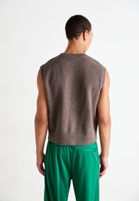 Gilet sans manches marron avec une encolure et un ourlet côtelés, associé à un pantalon vert ample avec une poche sur le côté.
