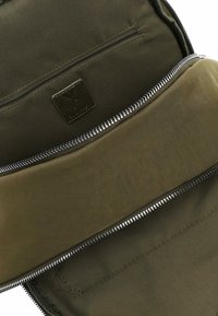 Otevřený olivově zelený batoh ukazující vnitřní kapsu na zip a textilní štítek uvnitř hlavního oddílu.