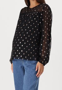 Blouse noire à manches transparentes et col rond, ornée de points métalliques dorés épars. Associée à un jean denim bleu clair.