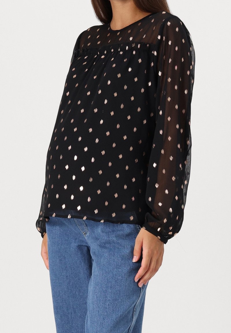 Blouse noire à manches transparentes et col rond, ornée de points métalliques dorés épars. Associée à un jean denim bleu clair.