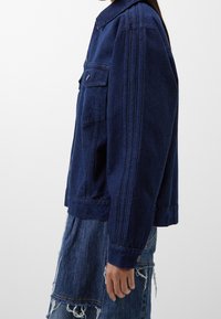 Tumma sininen farkkutakki, jossa on jäsennelty muotoilu, kaulus, kaksi etutaskua ja pystysuorat sauma- ja koristeyksityiskohdat.