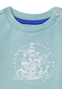 Jasnoniebieska koszulka bawełniana z grafiką białego zamku na plaży, z flagami, słońcem i narzędziami do budowy zamków z piasku. Żebrowany dekolt z detalem zatrzaskowym.