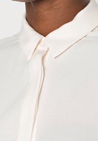 Blusa crema con colletto classico, realizzata in un tessuto leggero e morbido, con una discreta chiusura a bottoni sul davanti.
