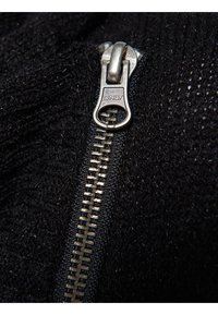 Zwart gestructureerd textiel met een metalen rits met een ronde trekker en merkteken, waarin het fijne weefsel en de details van de stof benadrukt worden.