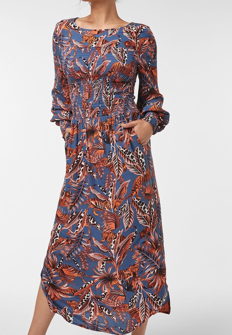 Femme portant une robe midi bleue à manches longues avec imprimé feuilles tropicales et animaux orange et noir, mains dans les poches latérales.