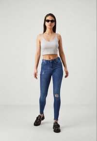 Skinny jeans in donkerblauwe denim met gescheurde accenten, gecombineerd met een grijze crop top en zwarte chunky schoenen. Zonnebrillen maken de look compleet.