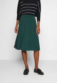 Grüner plissierter Rock mit glatter Textur und figurbetontem Bund. Kombiniert mit einem schwarzen Pullover und Loafers. Minimalistisches Design.