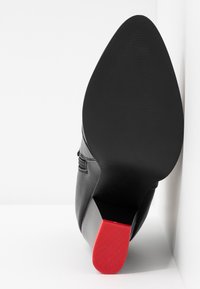 Schwarzer Leder-High-Heel mit spitzer Zehe und strukturiertem schwarzem Absatz, der ein markantes rotes Akzent am Absatz aufweist.