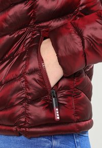 Rød pufferjakke med quiltet tekstur og en sidelomme. Har en sort lynlås med en rød trækstrop. Materialet ser glat og skinnende ud.
