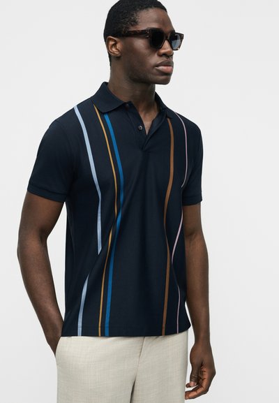 Homme portant des lunettes de soleil foncées, un polo bleu marine avec des rayures verticales bleues, marron et roses, et un pantalon beige clair sur un fond uni.