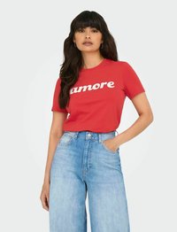 Rød bomulds t-shirt med hvid "amore" tekst, parret med lysblå vidde jeans. Modellen har langt mørkt hår og står med hænderne i lommerne.