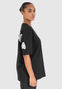 Schwarzes Baumwoll-T-Shirt mit kurzen Ärmeln, lockerer Passform und weißem Grafiktext auf dem linken Ärmel. Glatte Textur.
