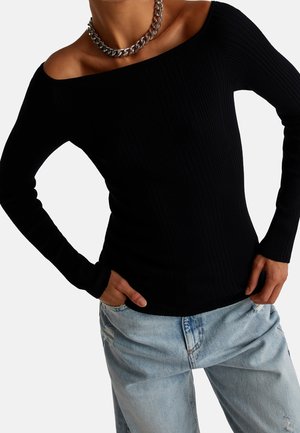 Femme portant un pull côtelé noir à épaules dénudées, un jean bleu clair déchiré, et un gros collier chaîne en argent, mains dans les poches du jean.