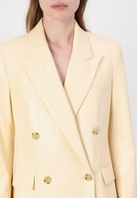 Blazer doppiopetto in pelle lucida di un giallo pallido con grandi bottoni dorati, revers a picco e una tasca sul petto.