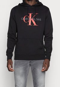 Sudadera negra hecha de una mezcla de algodón, con un estampado del logotipo de Calvin Klein en rojo y blanco en la parte frontal, con un bolsillo canguro y capucha con cuerda.