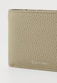 Portefeuille beige texturé avec une finition lisse, arborant le logo "Calvin Klein" en relief, des coutures subtiles et une forme rectangulaire élégante.