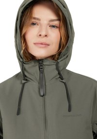 Chaqueta impermeable verde caqui con un cuello alto, cierre de cremallera, cordones ajustables y el logo de la marca en el pecho. Textura suave.