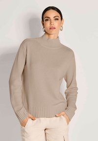 Frau mit dunklen Haaren, die hoop Earrings, einen beigen, gerippten Rollkragenpullover und helle Hosen trägt, mit den Händen in den Taschen.