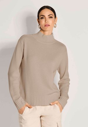 Frau mit dunklen Haaren, die hoop Earrings, einen beigen, gerippten Rollkragenpullover und helle Hosen trägt, mit den Händen in den Taschen.