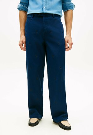 Mann trägt weite marineblaue Hose mit dezenten bestickten Mustern, hellblaues hochgekrempeltes Hemd und zweifarbige Loafers in Schwarz und Beige.