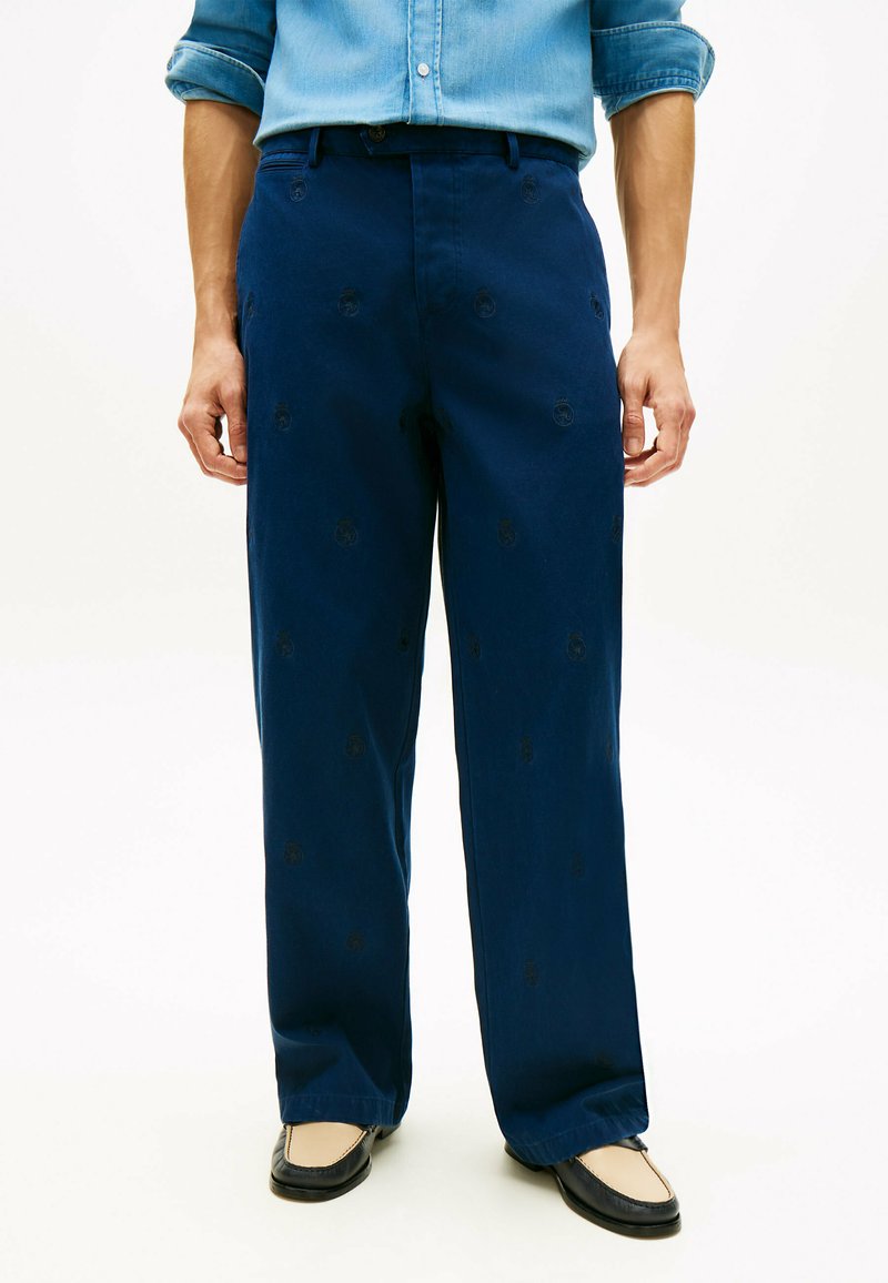 Homme portant un pantalon large bleu marine avec des motifs brodés subtils, une chemise bleu clair retroussée, et des mocassins bicolores noirs et beige.