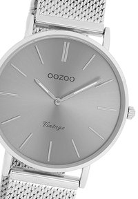 Silver metal wristwatch with a minimalist round face, featuring a brushed silver dial, thin hour markers, and a mesh strap. Brand name: OOZOO.