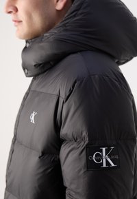 Schwarze Pufferjacke mit hohem Kragen, glänzender Textur und gesticktem Logo auf der Brust und dem Ärmel. Ausgestattet mit einem Reißverschluss.