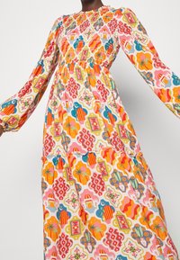 Maxikjole med lange ærmer, der har en smocket brystdel. Fremstillet af letvægtsstof, prydet med livlige, geometriske mønstre i rød, orange og blå.