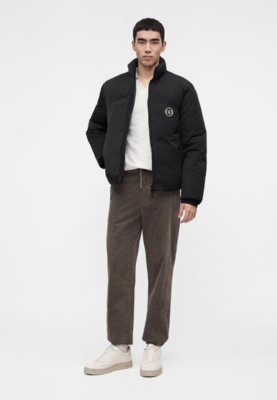 Homme portant une doudoune noire, une chemise blanche, un pantalon marron et des baskets blanches, debout avec une main dans la poche sur un fond uni.