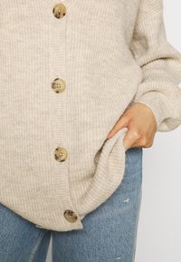 Beige pletený cardigan s ribkovanou texturou, s velkými světle hnědými knoflíky a boční kapsou, spárovaný s modrými roztrhanými džíny.