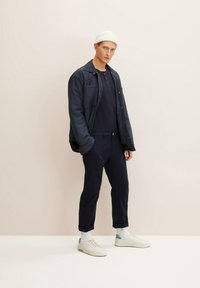 Hombre vestido con una chaqueta azul marino sobre una camiseta henley azul marino, combinada con pantalones azul marino y zapatillas blancas con detalles azules. Lleva un gorro de punto blanco en la cabeza.