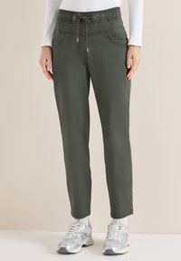 Personne portant un pantalon à cordon olive, un haut à manches longues blanc, des chaussettes blanches et des baskets argentées, debout devant un fond beige.