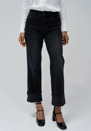 Jeans de mezclilla anchos negros con cintura alta, con puños enrollados y diseño de cinco bolsillos. Combinados con tacones negros con correa en el tobillo.