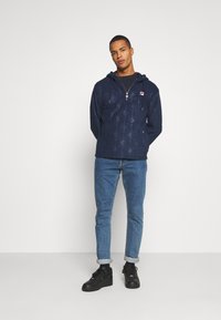 Felpa con zip di colore blu navy con un design a fantasia, caratterizzata da una tasca a marsupio frontale e coulisse, abbinata a jeans blu e sneakers nere.