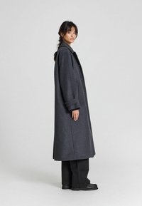 Cappotto lungo grigio scuro in lana con abbottonatura frontale, ampio colletto e vestibilità comoda. Presenta polsini arrotolati ed è indossato sopra pantaloni e scarpe neri.
