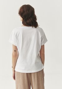 Bílá bavlněná trička s krátkými rukávy a volným střihem. Má kulatý výstřih a jednoduchý design, spárované s béžovými texturovanými kalhotami.