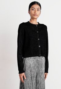 Black textured cardigan with a round neckline and six buttons, paired with a pleated gray skirt. The fabric appears soft and warm.