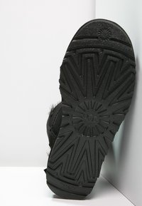 Czarna podeszwa buta UGG z głębokimi wzorami zygzaków i okręgów, przedstawiona w pionie na tle białego rogu.