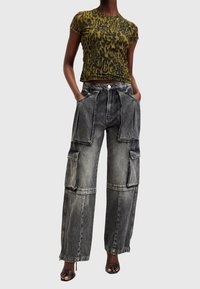 Person som bär en grön och svart leopardmönstrad kortärmad topp, grå vida cargobyxor med hög midja och stora fickor, samt svarta öppna klackskor.