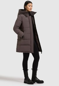 Puffer coat in muted taupe, featuring a high collar and vertical baffle design. Paired with black leggings and chunky black boots.