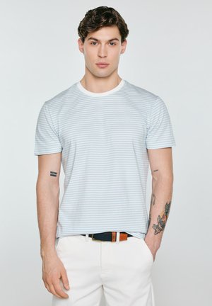 Mladý muž s hnědými vlasy, který nosí světle modré a bílé pruhované tričko a bílé kalhoty, stojí s jednou rukou v kapse.